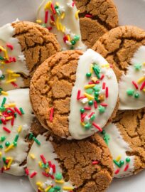 Close up of a ginger molasses cookie with a deep crackly top partially dipped in white chocolate and finished with festive Christmas sprinkles.