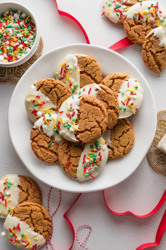 Plate of white chocolate dipped ginger molasses cookies, with extra sprinkles and red ribbons scattered nearby.