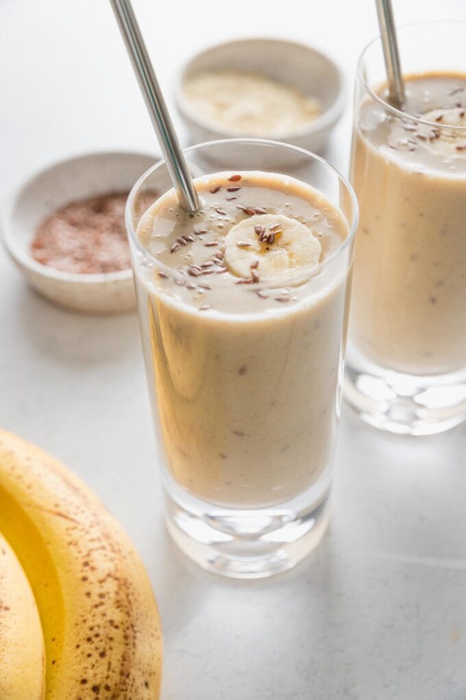Two banana protein shakes with an extra banana slice and flax seeds on top, served in tall clear glasses with metal straws.