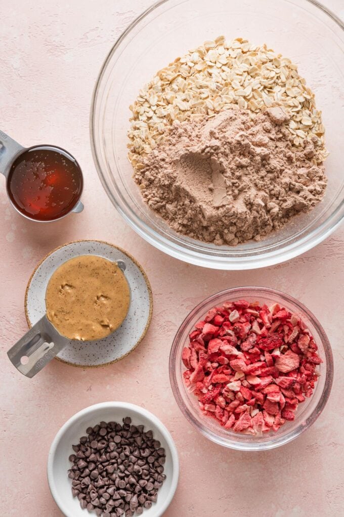 Overhead view of ingredients for protein bites: nut butter, honey, old-fashioned oats, chocolate protein powder, sea salt, mini chocolate chips, and freeze-dried strawberries.