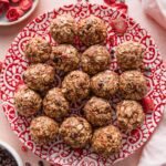 Small red and white patterned plate filled with chocolate strawberry protein bites, surrounded by extra mini chocolate chips and freeze-dried strawberries scattered on the countertop.