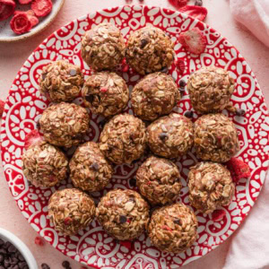 Small red and white patterned plate filled with chocolate strawberry protein bites, surrounded by extra mini chocolate chips and freeze-dried strawberries scattered on the countertop.