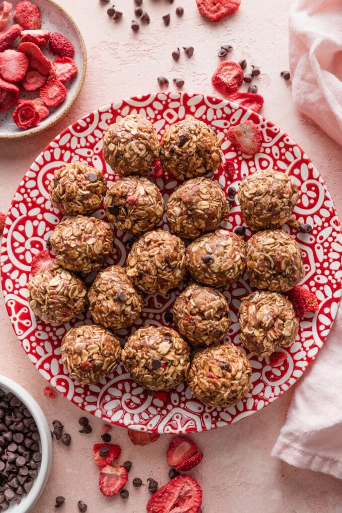 Small red and white patterned plate filled with chocolate strawberry protein bites, surrounded by extra mini chocolate chips and freeze-dried strawberries scattered on the countertop.
