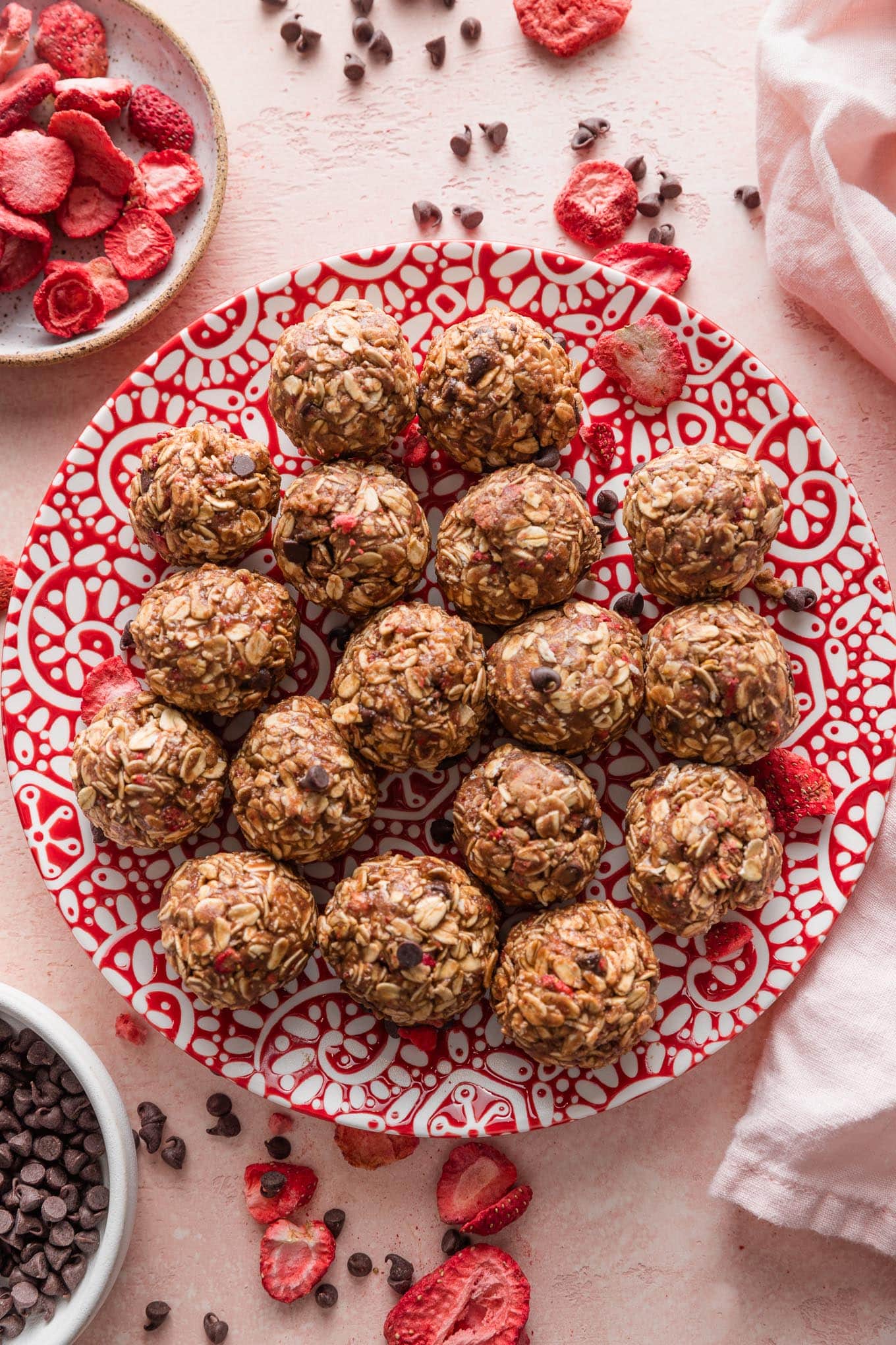 Small red and white patterned plate filled with chocolate strawberry protein bites, surrounded by extra mini chocolate chips and freeze-dried strawberries scattered on the countertop.