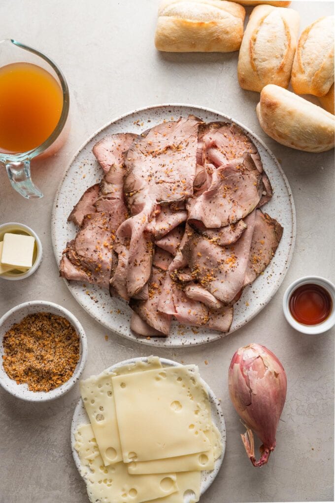 Overhead view of ingredients for simple French dips: deli roast beef, rolls, cheese, a shallot, steak seasoning, butter, beef broth, and sherry.