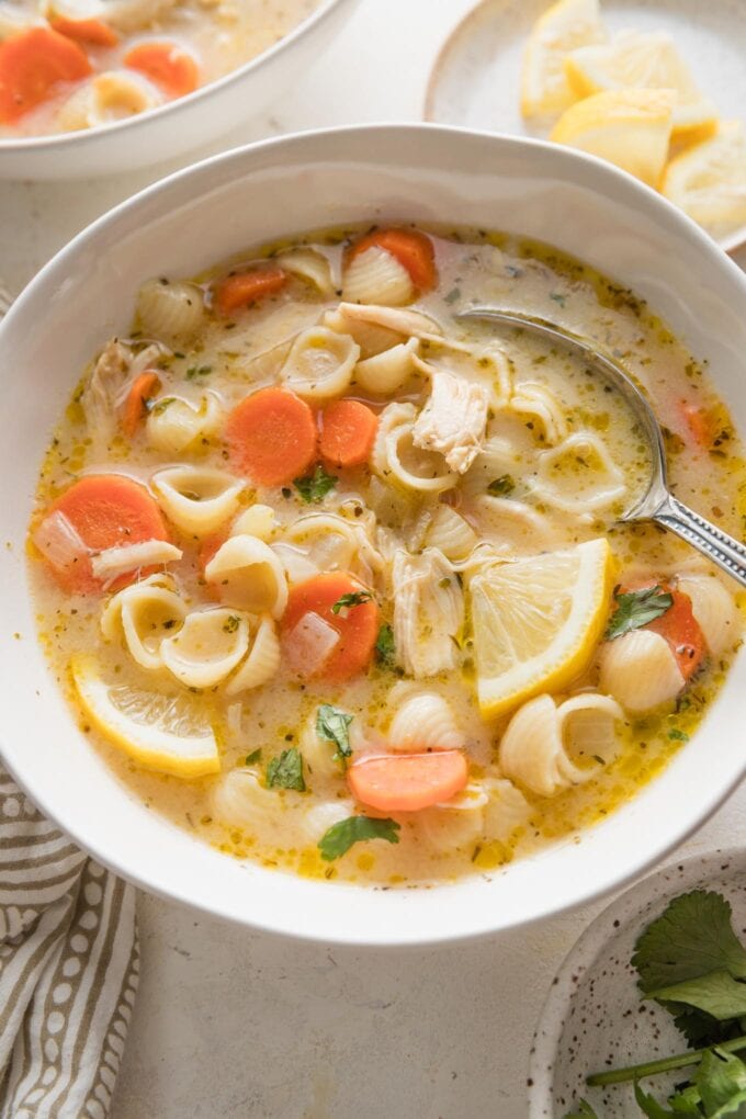 Angled view of a white ceramic bowl full of lemon chicken soup, with a spoon lifting out the first warm, brothy bite.