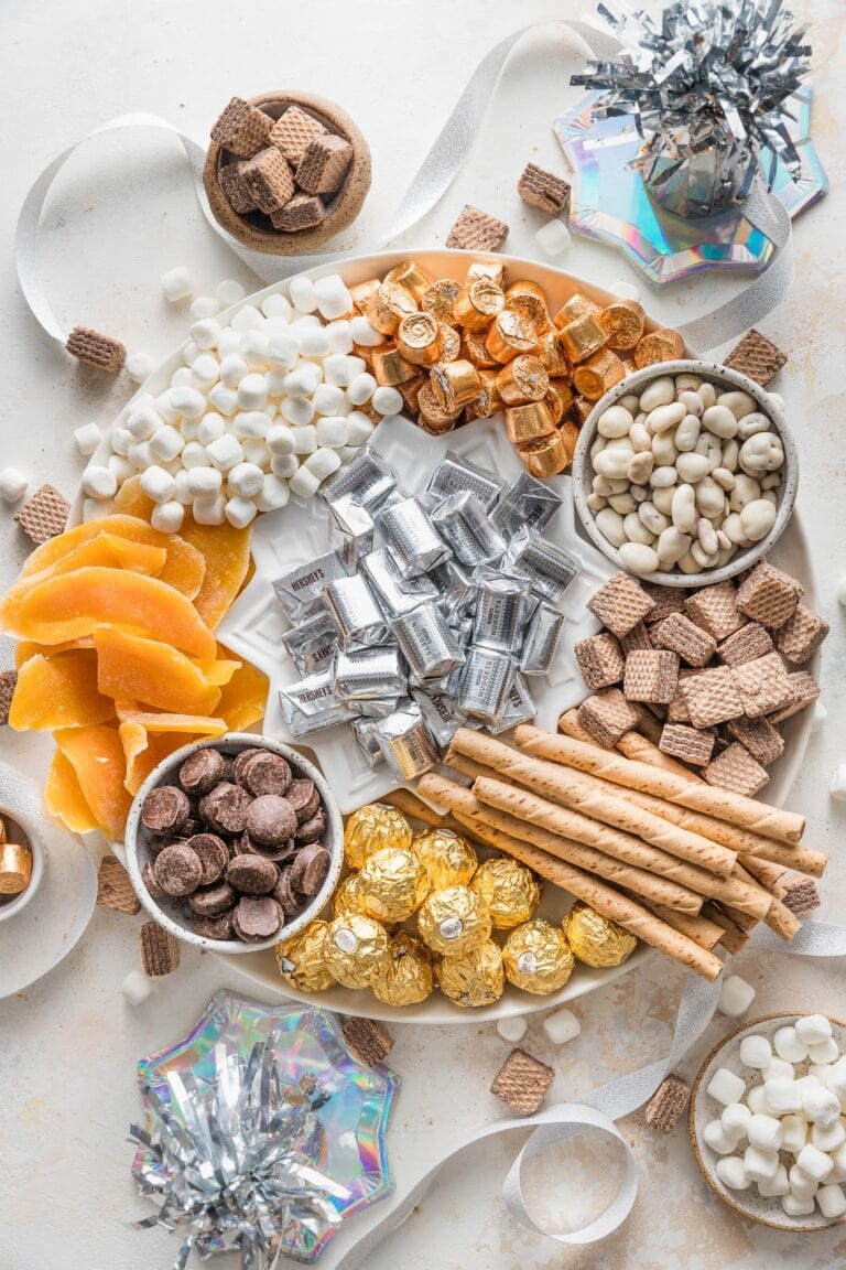 Large white platter filled with candy, dried fruit, and Pirouette cookies in silver, gold, and white tones, for celebrating New Year's Eve.