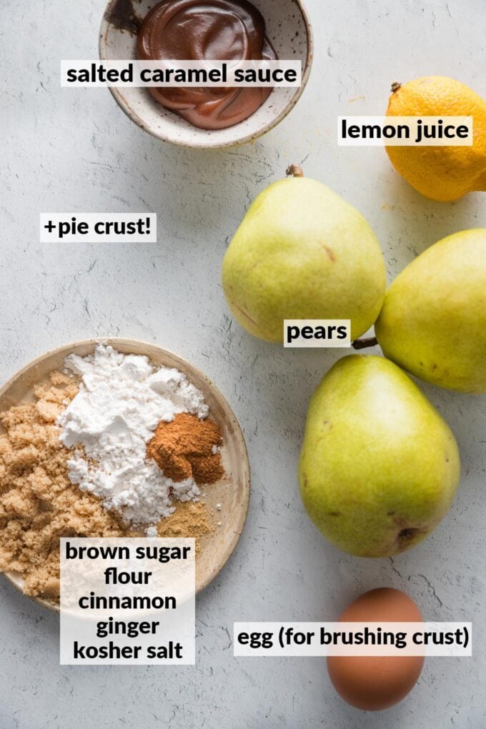 Overhead view of key ingredients for a pear caramel galette: pears, lemon, brown sugar, flour, cinnamon, ginger, kosher salt, salted caramel sauce, and an egg for brushing the crust.