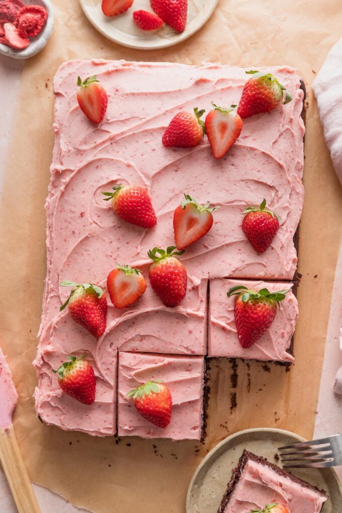 Overhead view of a chocolate sheet cake with strawberry frosting and fresh strawberries on top, removed from the pan and partially sliced into squares.