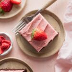 Small plate with a slice of chocolate sheet cake topped with strawberry frosting and a sliced fresh strawberry.