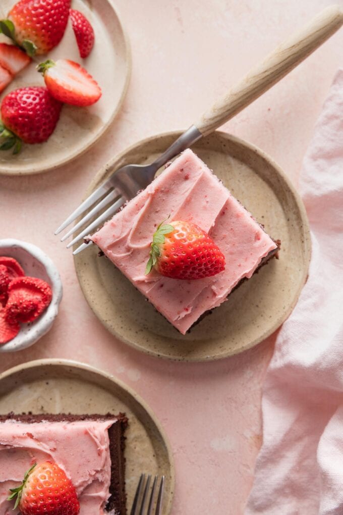Small plate with a slice of chocolate sheet cake topped with strawberry frosting and a sliced fresh strawberry.