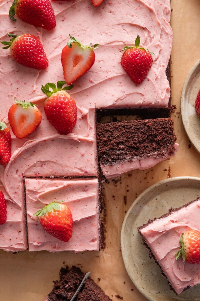 A large chocolate sheet cake topped with strawberry frosting and fresh sliced strawberries, removed from the pan and partially sliced to show some pieces with the frosting on top and others turned to show the rich, moist cake.