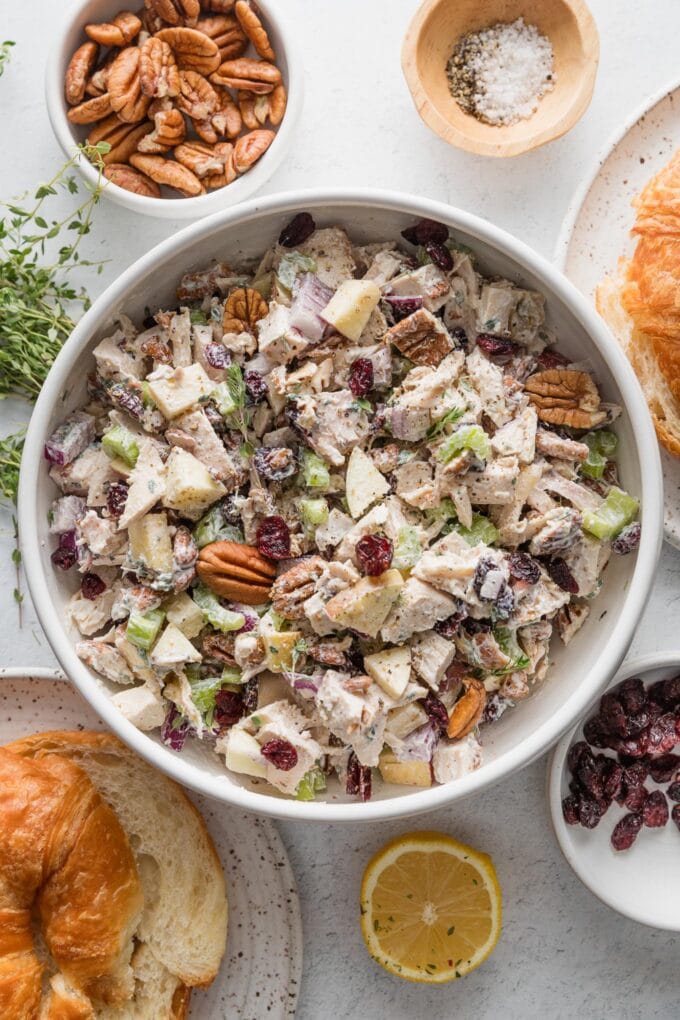 Overhead view of a large white ceramic bowl filled with cranberry pecan chicken salad, with extra pecans, dried cranberries, and croissants for serving neraby.