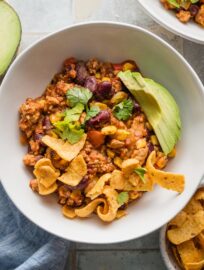 White bowl full of red chicken chili topped with avocado, corn chips, and fresh cilantro.