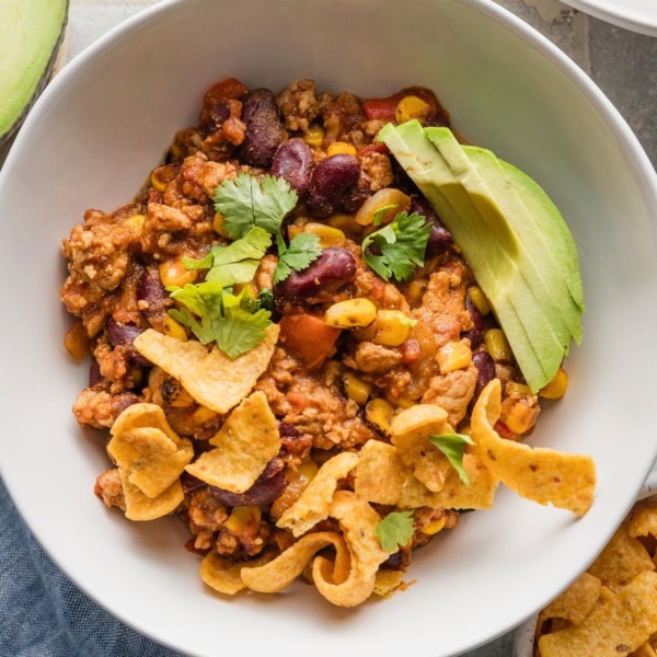 White bowl full of red chicken chili topped with avocado, corn chips, and fresh cilantro.