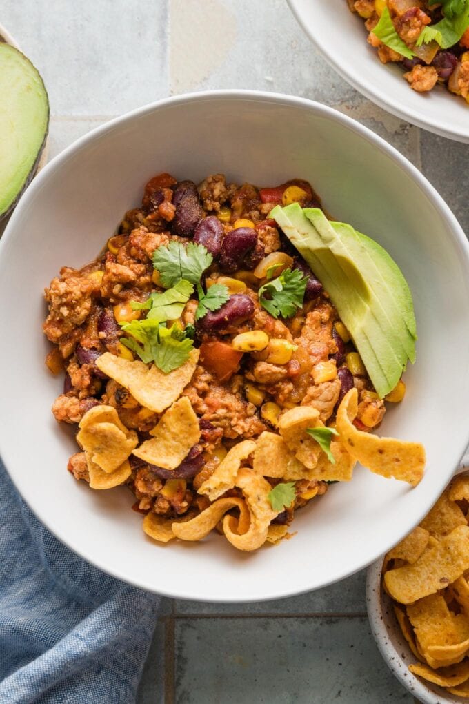 White bowl full of red chicken chili topped with avocado, corn chips, and fresh cilantro.
