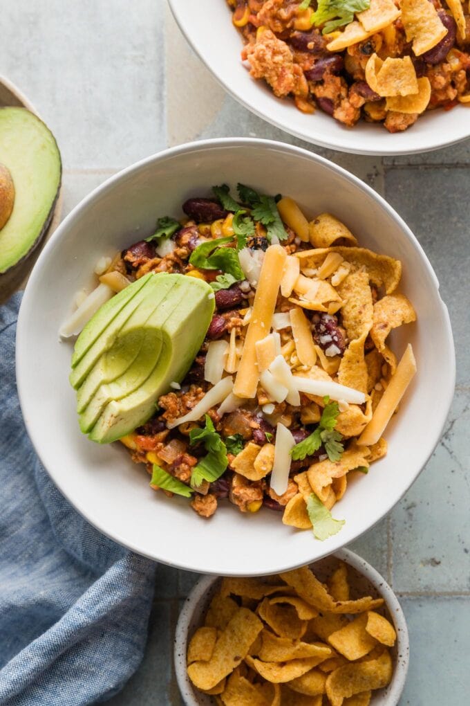 Bowls of ground chicken red chili with cheese, avocado, cilantro, and corn chips.