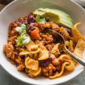Close up of a spoonful of red chicken chili made with ground chicken and a homemade spice blend, topped with corn chips and sliced avocado.