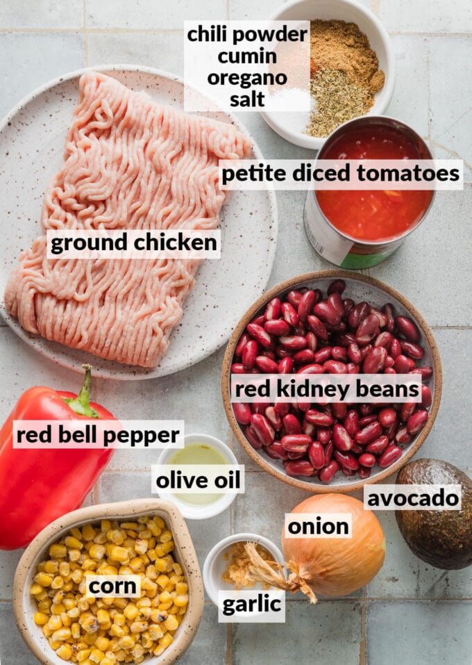 Labeled overhead photo of ingredients for red chicken chili: ground chicken, red kidney beans, frozen corn kernels, red bell pepper, onion, garlic, chili powder, cumin, oregano, and kosher salt.