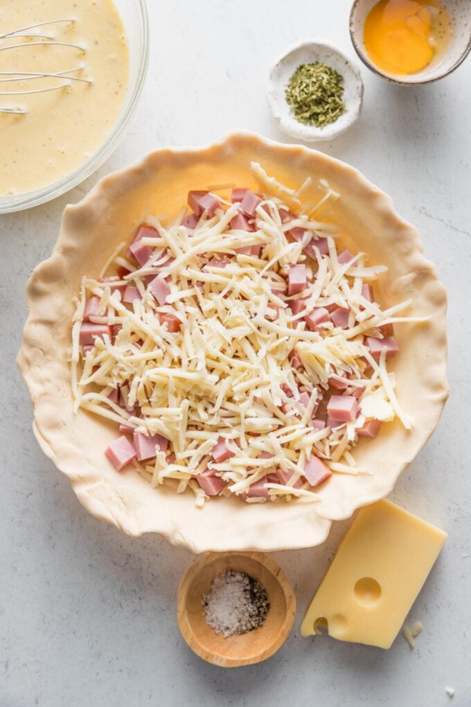 Pie crust pressed into a pie plate with the edges crimped and partially filled with cubed cooked ham and shredded cheese.