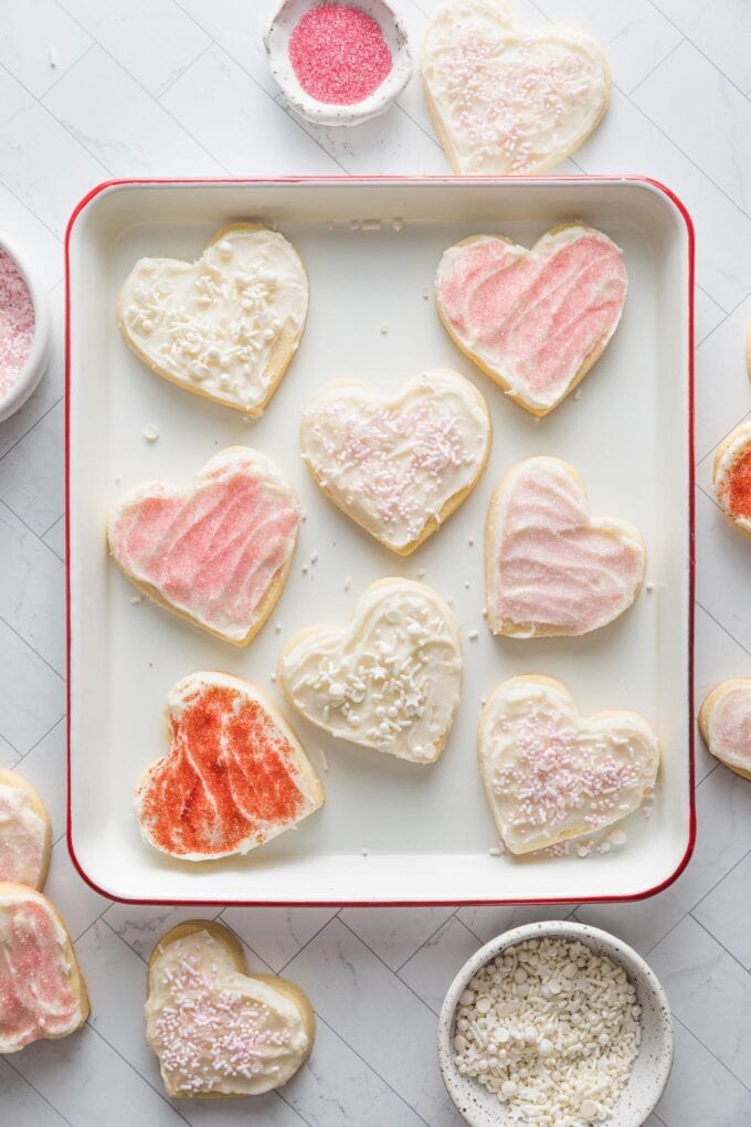 Tray filled with heart shaped cut out sugar cookies decorated with spreadable cream cheese cookie frosting and a mix of white, pink, and red sprinkles.