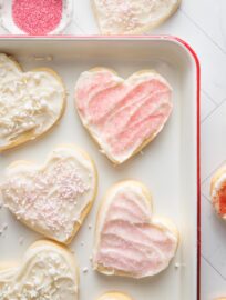 White enamel tray with heart shaped cut out sugar cookies and pink sprinkles in a small pinch bowl nearby.