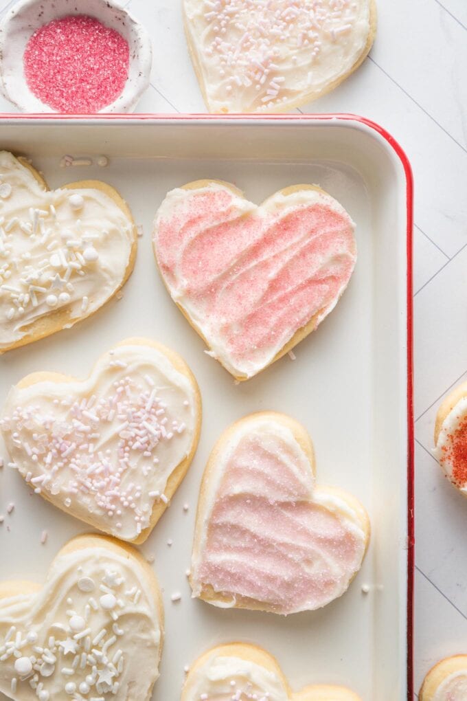 White enamel tray with heart shaped cut out sugar cookies and pink sprinkles in a small pinch bowl nearby.