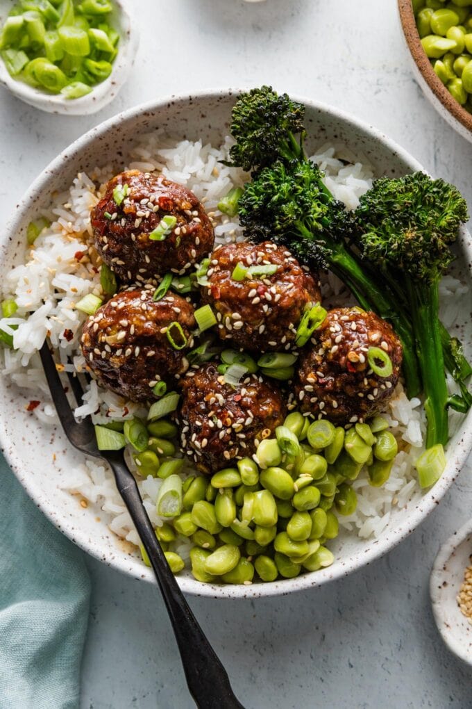 Bowl full of white rice topped with Asian-inspired ground beef meatballs in a sweet and sticky sauce topped with broccolini, shelled edamame, green onions, and toasted sesame seeds.