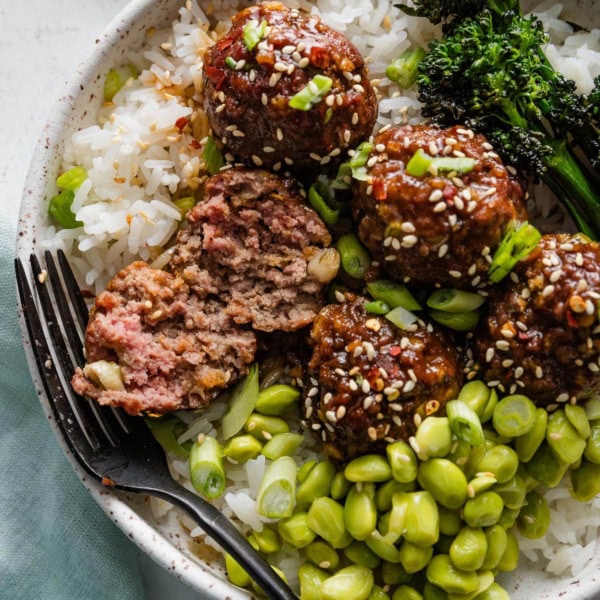Overhead view of sweet and sticky Asian meatballs served in a bowl with white rice, edamame, and broccolini, with one meatball split open to show the tender interior.