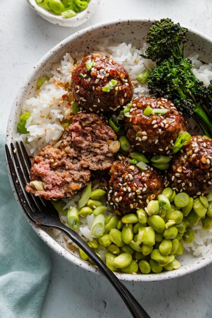 Overhead view of sweet and sticky Asian meatballs served in a bowl with white rice, edamame, and broccolini, with one meatball split open to show the tender interior.