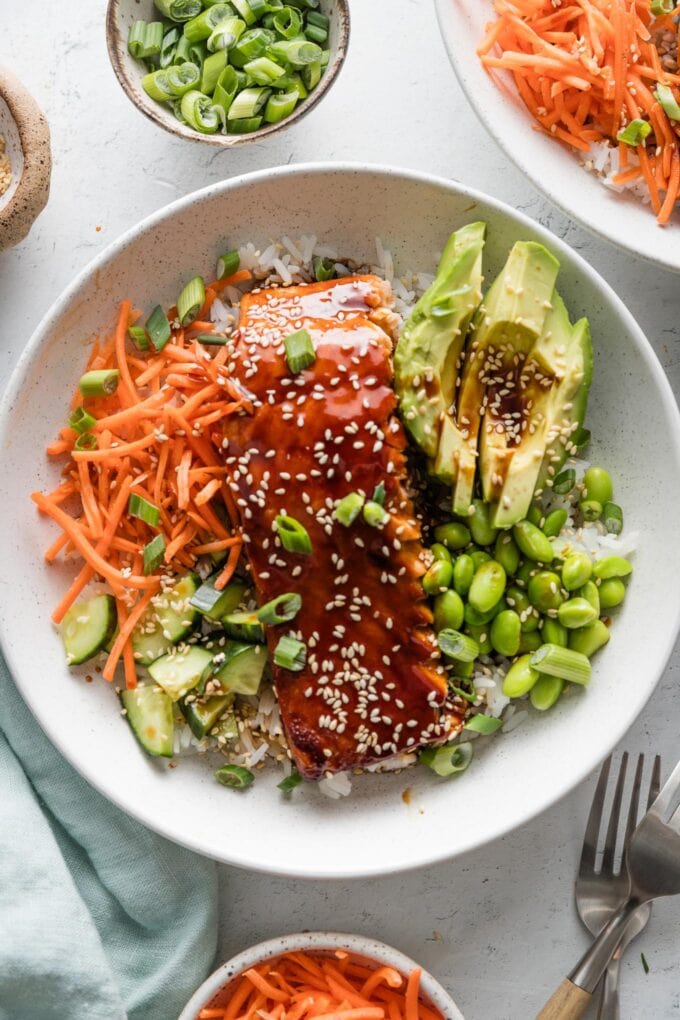 Overhead view of a teriyaki salmon bowl with avocado, edamame, carrots, cucumber, rice, sesame seeds and green onions.