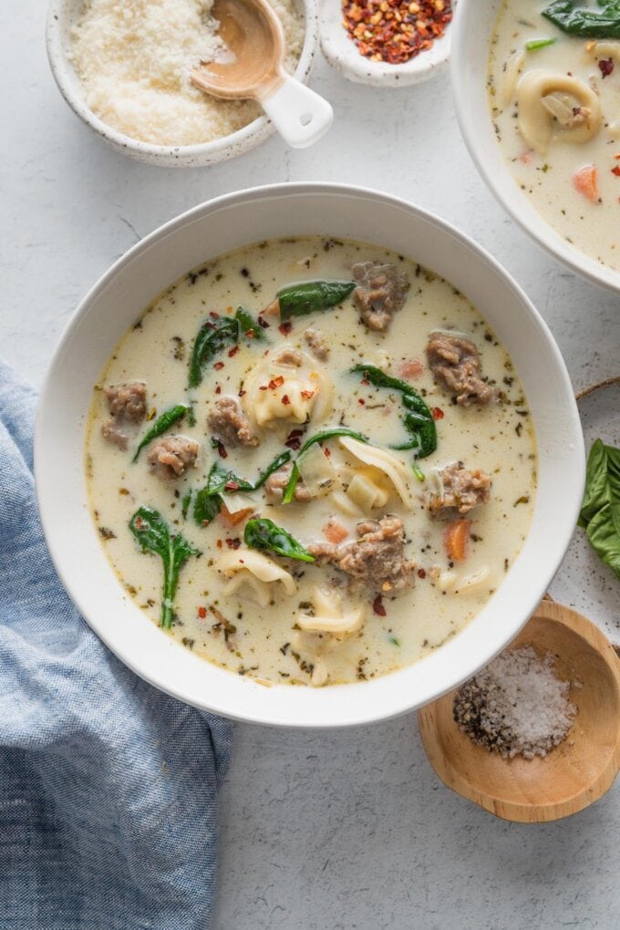 Countertop set with bowls of tortellini soup with cream broth and sausage, with Parmesan, red pepper, and extra basil leaves nearby to top bowls.