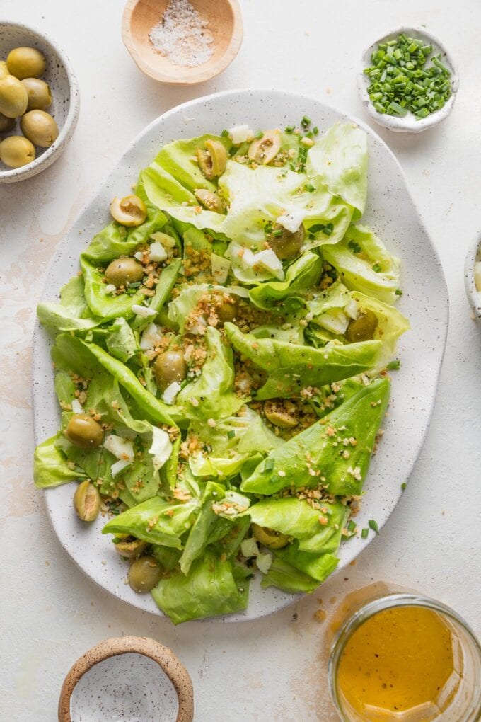 Countertop set with a large platter of butter lettuce salad with Castelvetrano olives, surrounded by extra toppings: chives, Manchego cheese, dressing.