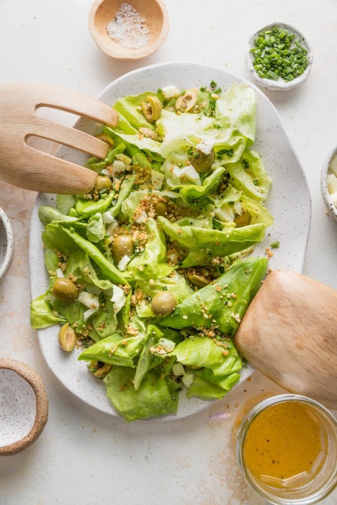 Salad tongs resting on a platter filled with a Bibb lettuce salad with Manchego, Castelvetrano olives, and toasted Panko.