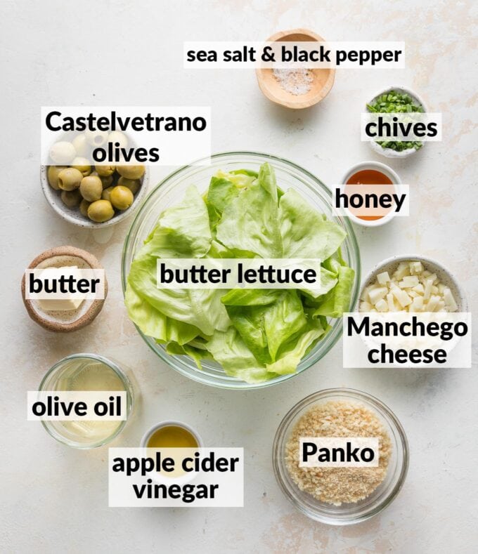 Labeled overhead image of washed butter lettuce leaves, pitted Castelvetrano olives, crumbled Manchego cheese, Panko, butter, chives, honey, apple cider vinegar, olive oil, salt, and pepper, all in prep bowls and ready to assemble into a salad.