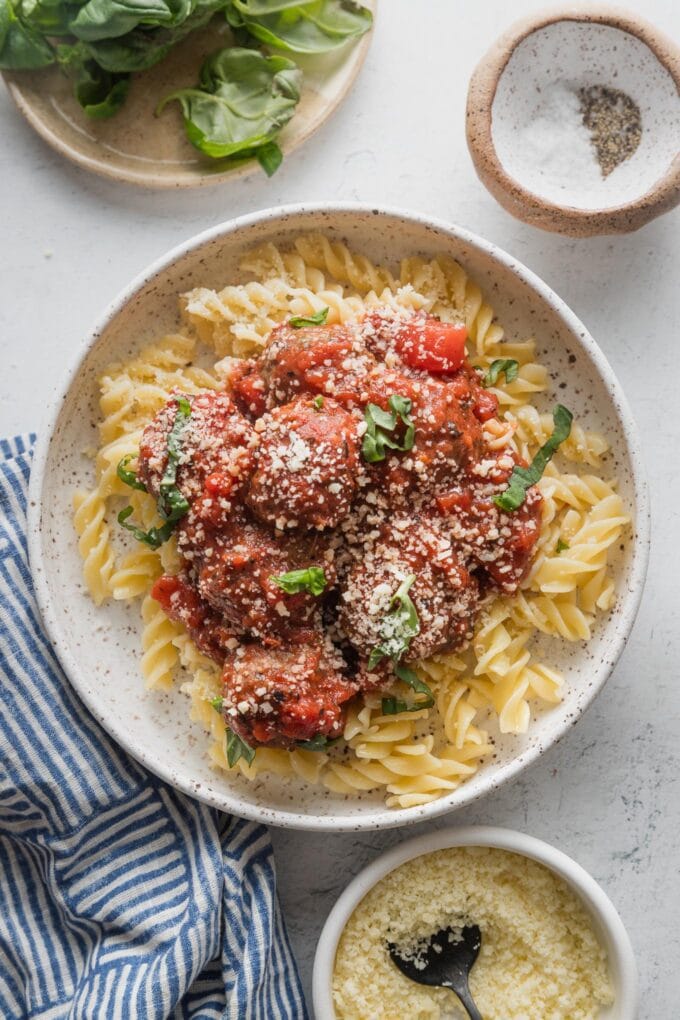 Countertop set with a bowl of meatballs marinara, with extra fresh basil, Parmesan, salt and pepper scattered nearby.