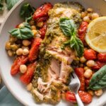 Bowl of pesto salmon baked with cherry tomatoes and chickpeas and topped with a squeeze of fresh lemon juice and fresh basil, with part of the salmon flaked off with a fork.