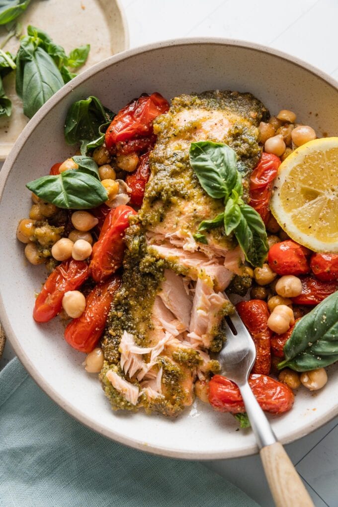 Bowl of pesto salmon baked with cherry tomatoes and chickpeas and topped with a squeeze of fresh lemon juice and fresh basil, with part of the salmon flaked off with a fork.