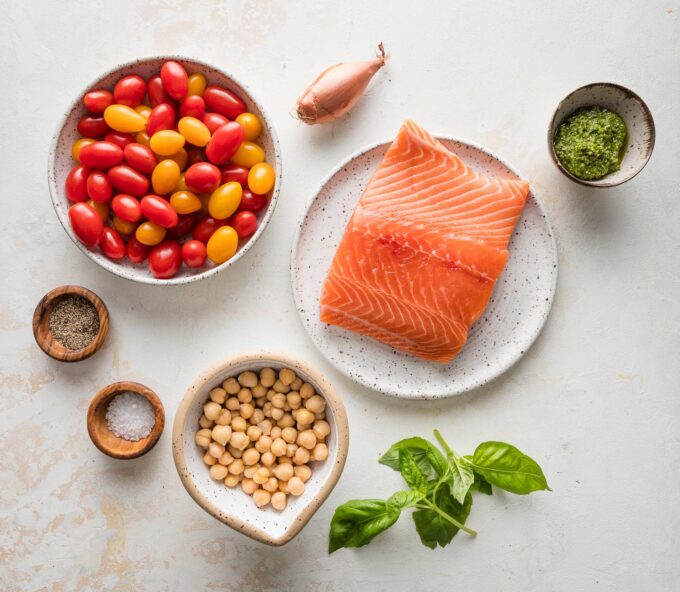 Overhead photo of a large salmon filet, cherry tomatoes, chickpeas, shallot, basil, pesto, salt, and pepper.