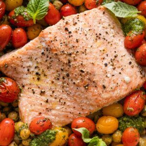 Close up of oven roasted salmon with pesto, tomatoes, chickpeas, and shallot in a shallow enameled roasting pan.