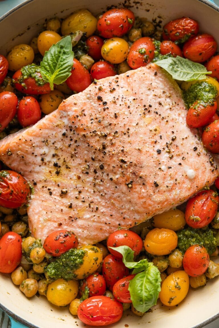 Close up of oven roasted salmon with pesto, tomatoes, chickpeas, and shallot in a shallow enameled roasting pan.