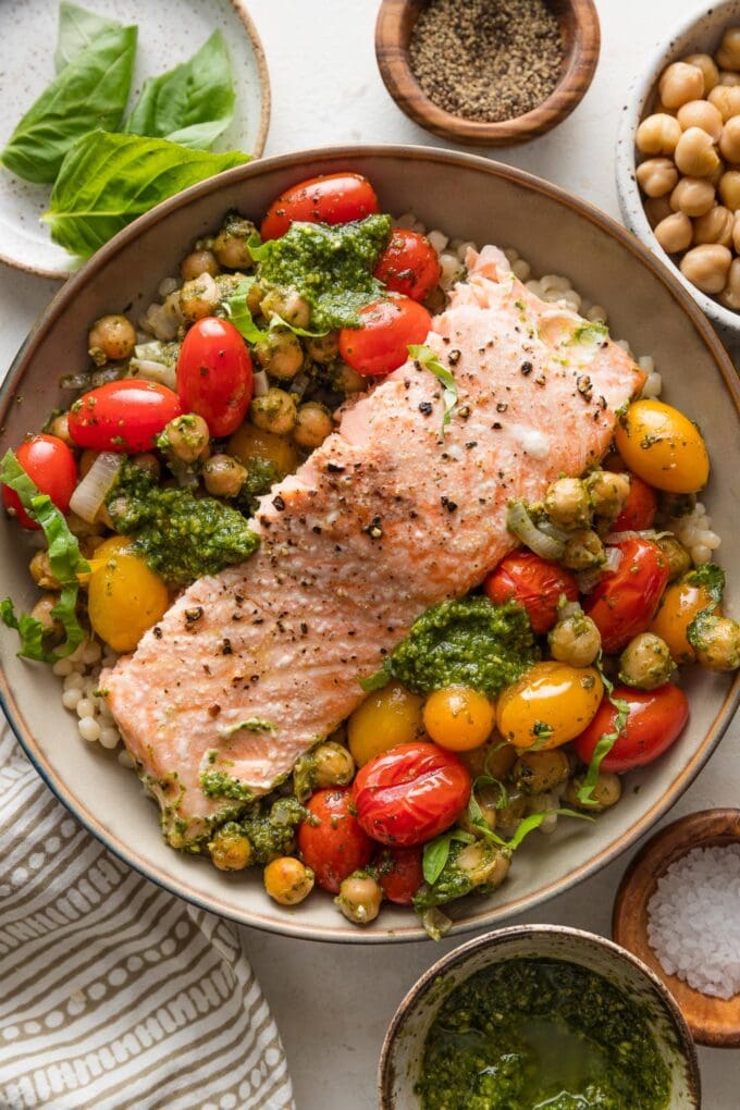 Bowl full of oven roasted salmon, cherry tomatoes, chickpeas, and shallot with basil pesto, served over pearl couscous.