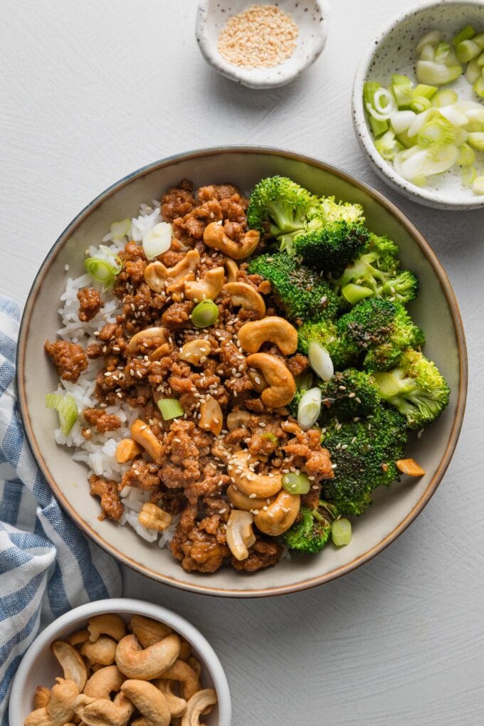 Bowl of sesame cashew chicken served over rice with extra cashews, slivered green onions, and toasted sesame seeds nearby.