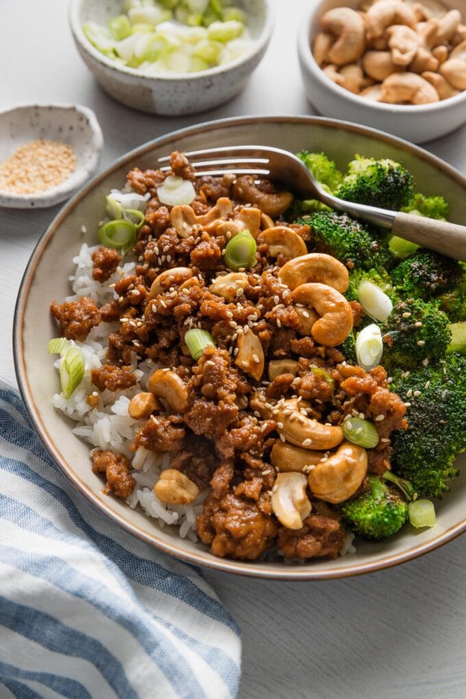 Close up of a fork stuck into a bowl full of toasted sesame cashew chicken.