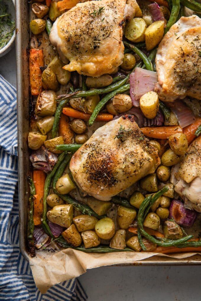 Overhead view of a large rimmed sheet pan with roasted chicken thighs and veggies seasoned with Herbes de Provence.