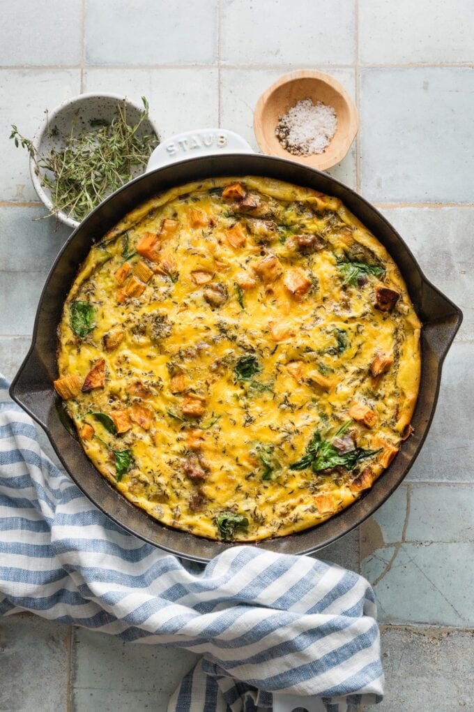 Overhead view of a counter with a cooling sweet potato sausage frittata with extra kosher salt, black pepper, and fresh thyme scattered nearby.