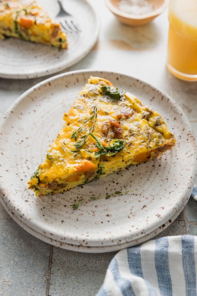 Angled view of a small plate with a slice of sweet potato and sausage frittata, with extra slices and a small glass of orange juice in the background.
