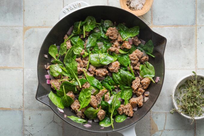 Small cast iron skillet filled with crumbled browned sausage, red onion, spinach, and garlic.