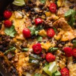 Close up of an enchilada skillet made with black beans and ground beef, topped with tomatoes, lime wedges, and cilantro leaves.