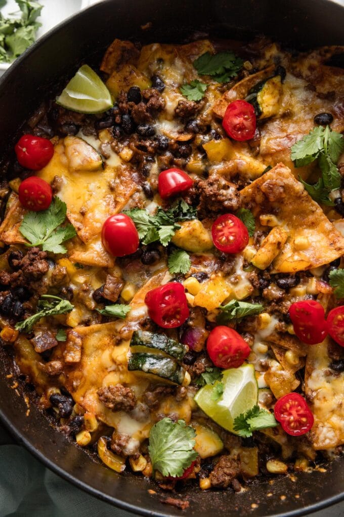 Close up of an enchilada skillet made with black beans and ground beef, topped with tomatoes, lime wedges, and cilantro leaves.